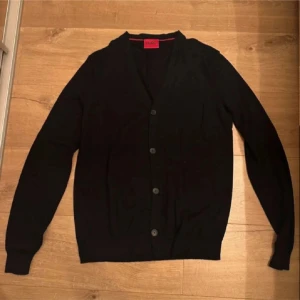 Svart kofta från Hugo Boss - Snygg svart kofta från Hugo Boss med knappar framtill. Perfekt för lager-på-lager och passar till många olika stilar. Den har en klassisk design med långa ärmar och ribbade muddar.