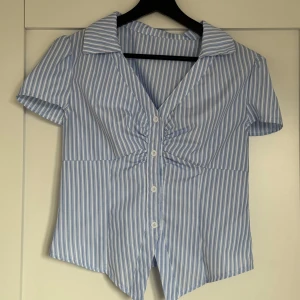 Blåvit randig skjorta med v-ringning - Kortärmad skjorta med blå och vita ränder, djup v-ringning och snygg rynkdetalj framtill. Skjortan har klassisk krage, knappar hela vägen och en unik spetsig nederkant både fram och bak. Perfekt för dig som gillar en fräsch och stilren look.