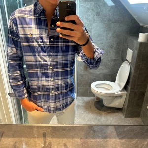 Blårutig skjorta från Ralph Lauren - Säljer en klassisk blårutig skjorta från Ralph Lauren med button-down krage och broderad logga på bröstet. Skjortan har lång ärm, normal passform och är tillverkad i mjuk bomull. Perfekt för dig som gillar stilren och tidlös look.