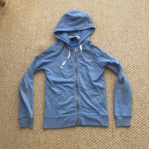 Blå hoodie från Gant med dragkedja - Snygg ljusblå hoodie från Gant i storlek XS. Tröjan har huva med vita snören, hel dragkedja och två fickor framtill. Tillverkad i mjuk bomull, perfekt för chill dagar. Gant-logga broderad på bröstet för en clean look.