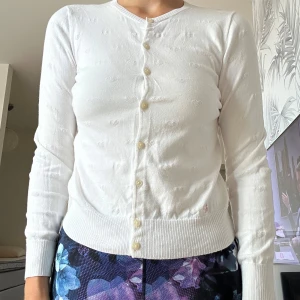 Vit kofta med hjärtmönster och blommor POLO - Säljer en söt vit kofta med små invävda hjärtan och en broderad blomdetalj vid nederkanten. Koftan har rund hals, långa ärmar och knäpps med pärlemorskimrande knappar framtill. Perfekt för dig som gillar gulliga detaljer och mjuka material.storlek 16 år 