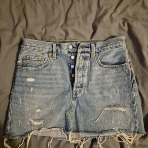 Levi's ljusblå jeanskjol med slits - Säljer en snygg ljusblå jeanskjol från Levi's med slitna detaljer och fransig kant. Kjolen har klassiska fickor fram och bak, knappstängning och bälteshällor. Perfekt för dig som gillar en chill och trendig streetstil.