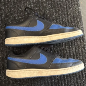 Nike Court Vision sneakers blå/svarta - Säljer ett par Nike Court Vision sneakers i svart och blått skinn med klassisk vit sula. Skorna har rund tå, snörning och den ikoniska Nike-swooshen i blått på sidorna. Perfekta för dig som gillar sportig och stilren vibe.