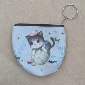 Söt plånbok med kattmotiv och dragkedja - Liten plånbok med ljusblå bakgrund och ett gulligt tryck av en tecknad kattunge på ett garnnystan. Plånboken har dragkedja upptill och en metallring för nycklar. Perfekt för mynt, kort eller småprylar. Materialet känns som syntet och ytan är lätt strukturerad.