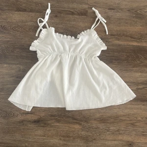 Vit linnetopp med volang från 157 - Supersöt vit linnetopp från 157 med tunna axelband som knyts och volangdetaljer längs urringningen. Toppen har en lätt rynkad struktur och är luftig med en flowy passform, perfekt för varma dagar. Elastisk midja för extra komfort och snygg siluett. Skriv för egna bilder💖
