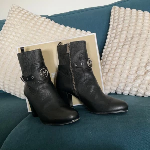 Svarta boots från Michael Kors - Snygga svarta boots från Michael Kors i skinn med rund tå och hög klack. Dekorativa remmar med silverfärgade MK-loggor och dragkedja på sidan. Perfekta för dig som vill ha en stilren och edgy look.