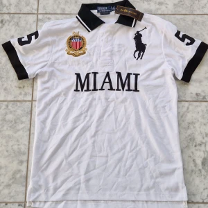 Vit Polo Ralph Lauren Miami t-shirt - Vit t-shirt från Polo Ralph Lauren med svart krage och svarta detaljer på ärmarna. Stor broderad logga och texten MIAMI på bröstet, samt siffran 5 på både ärm och rygg. Klassisk polodesign med korta ärmar och broderade emblem. Materialet är 100% exklusivt bomull. Riktigt trendig, passar till alla stiler. Storlek S, passar även XS (se måtten) Hör av dig vid minsta fråga eller prisförslag! 🤍