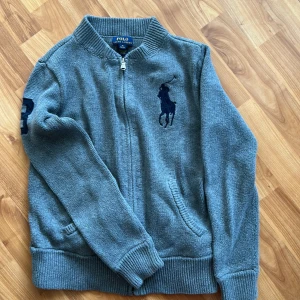 Barn- Grå stickad kofta Polo Ralph Lauren - Barn-tröja - Grå stickad kofta från Polo Ralph Lauren med dragkedja framtill, ribbade muddar och krage, två fickor framtill . Fint skick