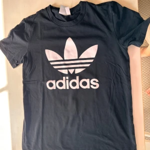 Svart Adidas t-shirt med logga - Svart t-shirt från Adidas med stor vit trefoil-logga och text på framsidan. Klassisk rund hals och korta ärmar. Tillverkad i mjuk bomull som är skön mot huden. Perfekt för dig som gillar sportiga och stilrena plagg med tydlig branding.