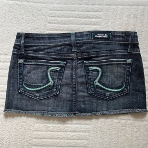 Rock & Republic jeanskjol med fickor - Supersnygg mörkblå jeanskjol från Rock & Republic med fransad kant och coola bakfickor med turkos broderi. Klassisk femficksmodell, låg midja och silverfärgade knappar. Perfekt för dig som gillar streetstyle och denim.