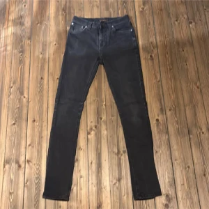 Svarta jeans från Nudie Jeans - Säljer ett par svarta skinny jeans från Nudie Jeans med klassisk femficksdesign och diskret logga på bakfickan. Jeansen har smal passform och är tillverkade i stretchig bomullsdenim för en bekväm känsla. Perfekta för dig som gillar en stilren look.