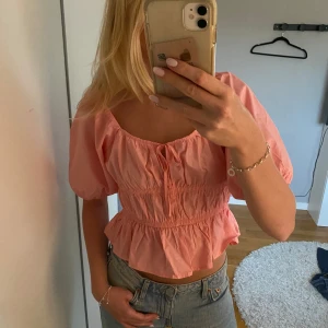 Rosa blus med puffärm från H&M - Supersöt ljusrosa blus från H&M med puffiga korta ärmar, smockad midja och volang nedtill. Blusen har en söt knytning framtill och är croppad, perfekt till jeans eller kjol. Tillverkad i mjuk bomull som känns lätt och luftig. Aldrig använt med prislapp😍
