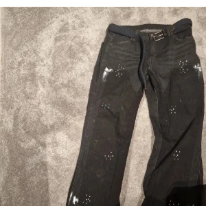 Svarta denim tears - Unika svarta jeans med vita nitar och detaljerade prickmönster över hela benen. Råa, avklippta benslut för en edgy look. Byxorna har bälte i midjan och är tillverkade i kraftigt jeanstyg. Perfekta för dig som vill sticka ut.