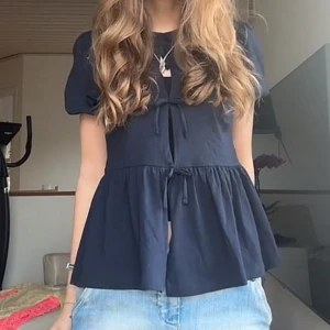 Svart blus med knyt och volang - Supersöt svart blus med korta puffärmar, två knyt framtill och volang nedtill. Blusen har en luftig och flowy passform som ger en avslappnad men ändå trendig look. Perfekt att styla med jeans eller kjol för en cool vardagsoutfit.