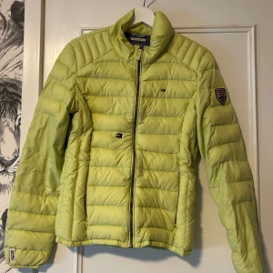 Gul pufferjacka från American Brand - Gul pufferjacka från American Brand med broderad badge på ärmen och dragkedja framtill. Jackan har quiltad design, hög krage och klassisk passform. Perfekt för dig som vill sticka ut med färg. Materialet är syntet och jackan är långärmad.
