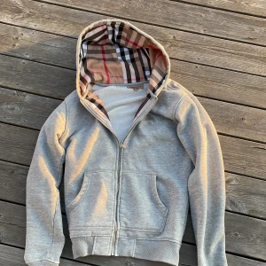 Grå Burberry hoodie - Säljer en grå hoodie från Burberry med klassisk rutig insida på huvan och broderad logga på bröstet. Tröjan har dragkedja, två fickor fram och ribbade muddar. Perfekt för dig som vill ha en clean look med lyxig touch.