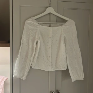 Vit blus med broderade detaljer från ONLY - Superfin vit blus från ONLY med broderade blommor och knappar framtill. Blusen har offshoulder-modell, långa ärmar och smockad rygg för extra bekväm passform. Perfekt för dig som gillar romantisk och luftig stil.