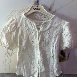 Vit blus med broderade hål från Zara - Superfin vit blus från Zara med broderade hål och puffiga korta ärmar. Blusen har knappar framtill och är gjord i ett lätt, krinklat bomullstyg som känns luftig och somrig. Perfekt för dig som gillar romantiska detaljer och en chill vibe.