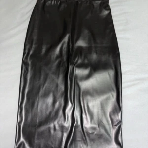 Svart lång kjol i skinn från ASOS - Säljer en svart lång kjol från ASOS DESIGN i snyggt, glansigt skinnimitation. Kjolen har en rak siluett och en slits bak för extra edge. Perfekt för dig som gillar minimalistisk och cool stil. Elastisk midja för skön passform.