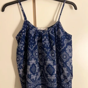 Blå mönstrad linnetopp från 157 - Supersnygg blå linnetopp från 157 med tunna axelband och vitt paisley-inspirerat mönster. Toppen har en loose passform och är perfekt för varma dagar. Materialet känns lätt och skönt mot huden, och färgen är riktigt fräsch och poppar till outfiten.