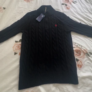 Svart stickad polotröja Polo Ralph Lauren - Helt ny svart kabelstickad polotröja från Polo Ralph Lauren med halv dragkedja och röd broderad logga på bröstet. Tröjan har ribbade muddar, hög krage och är långärmad. Perfekt för dig som gillar klassisk och stilren look.