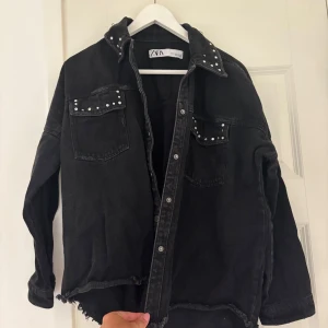 Svart jeansjacka med nitar från Zara - Cool svart jeansjacka från Zara med nitar på kragen och fickorna. Jackan har råa kanter nertill, knappar framtill och två bröstfickor. Perfekt för dig som vill ha en edgy vibe till din outfit.