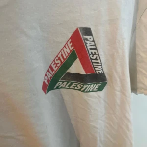 Vit Palestine t-shirt med tryck - Vit t-shirt med stort grafiskt Palestine-tryck i rött, svart och grönt både fram och bak. Klassisk rund hals och korta ärmar. Skön och avslappnad passform, perfekt för dig som vill sticka ut med ett statement-plagg.