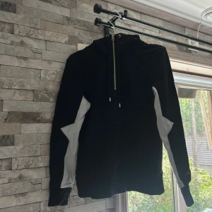 Svart hoodie från Elly Pistol med dragkedja - Cool svart hoodie från Elly Pistol i storlek S med vita detaljer på ärmarna och stor vit logga på ryggen. Tröjan har huva med snörning och en snygg dragkedja framtill. Materialet är mjukt och känns som bomull eller polyester.