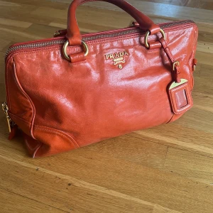 Orange handväska från PRADA i skinn - Säljer en snygg orange handväska från Prada i äkta skinn med guldiga metalldetaljer och dragkedja. Väskan har dubbla handtag, avtagbar axelrem och klassisk Prada-logga framtill. Perfekt för dig som vill sticka ut med en färgstark accessoar.