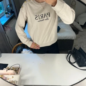 Beige sweatshirt med guldknappar, H&M - Beige sweatshirt från H&M med svart text 'PARIS Place Vendôme' framtill. Tröjan har rund halsringning, långa ärmar och fyra dekorativa guldknappar på ena axeln. Mjuk insida och relaxed fit, perfekt för en chill och snygg look.