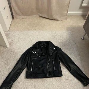 Svart skinnjacka från Zara - Snygg svart skinnjacka från Zara med klassisk bikerstil. Jackan har silvriga dragkedjor, fickor med lock och coola detaljer vid axlarna. Croppad modell med lång ärm och markerad krage. Perfekt för dig som vill ha en edgy look.