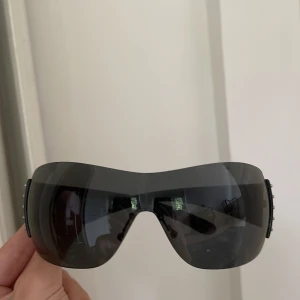 Svarta Prada solglasögon med nitar - Säljer ett par svarta Prada solglasögon med futuristisk shield-form och breda skalmar dekorerade med silvriga nitar och logga. Glasögonen har mörka linser och en y2k vibe, perfekta för dig som vill sticka ut. Köpta för några år sedan second hand för 2000 kr.