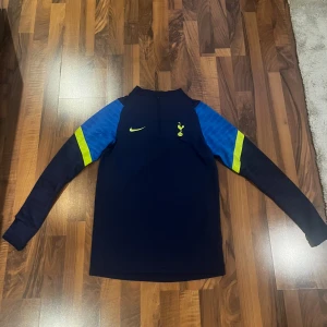 Tottenham långärmad träningströja Nike - Sällsynt mörkblå långärmad Tottenham Hotspur träningströja från Nike med halv dragkedja. Ärmarna har blå partier och gula detaljer. Tröjan har klubbens emblem och Nike-logga i gult på bröstet. Tillverkad i funktionsmaterial som andas.