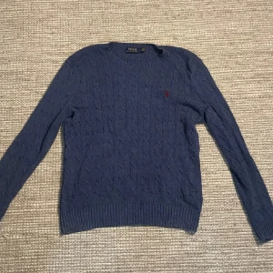Blå kabelstickad tröja Polo Ralph Lauren - Kabelstickad blå tröja från Polo Ralph Lauren med rund halsringning och klassisk röd broderad logga på bröstet. Tröjan är långärmad och har ribbade muddar vid ärmslut och nederkant. Perfekt för dig som gillar preppy och stilren look. Köpt nyss men passade tyvärr inte så alldrig använd.