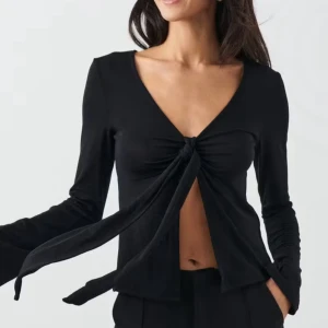 Svart omlottopp med knyt från Glamorous - Svart omlottopp från Glamorous med lång ärm och djup v-ringning. Toppen har en snygg knytning framtill som ger en öppen look vid midjan. Materialet är mjukt och stretchigt, perfekt för att skapa en cool och trendig outfit.