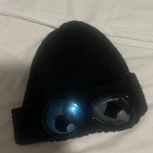 Svart C.P. Company goggle-mössa - Svart ribbstickad mössa från C.P. Company med ikoniska blåtonade goggle-linser framtill. Mössan har en uppvikt kant och är tillverkad i mjuk ull, perfekt för att ge en cool och unik look under kalla dagar.