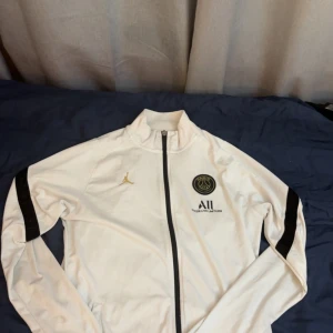 Vit PSG x Jordan  - Snygg vit vindjacka från PSG och Jordan med svarta detaljer på ärmarna och dragkedja. Koftan har guldiga loggor på bröstet och hög krage. Perfekt för dig som gillar sportig stil och vill sticka ut med exklusiva samarbeten.
