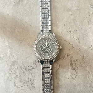 Glittrig silverklocka med stenar - Säljer en riktigt snygg klocka i silverfärg med massor av glittrande stenar både på urtavlan och armbandet. Urtavlan har romerska siffror och hela klockan har en lyxig vibe. Perfekt för dig som gillar att sticka ut och vill ha något som blänker ordentligt. Obs inget batteri har bara använts som accessoar