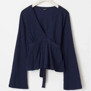 Mörkblå omlottopp med knyt  - Supersnygg mörkblå omlottopp från Ginatricot i storlek XS. Helt ny med prislapp kvar! Toppen har v-ringning, långa ärmar och ett knytband i midjan som ger en schysst siluett. Materialet är mjukt och lätt strukturerat, perfekt för dig som gillar stilrena och bekväma plagg.