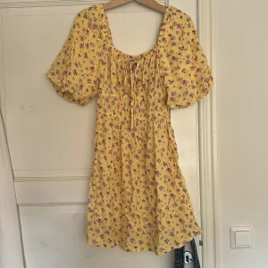 Gul blommig kortklänning med puffärm - Superfin gul kortklänning med lila och rosa blommor, puffiga korta ärmar och smock i midjan. Söt snörning framtill och mjukt, luftigt material som känns perfekt för varma dagar. Klänningen har en feminin och avslappnad vibe med volym i ärmarna.