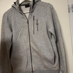Grå hoodie från Sail Racing med dragkedja - Säljer en grå hoodie från Sail Racing med hel dragkedja och huva. Tröjan har en snygg bröstficka med dragkedja och logga på vänster sida. Mjuk insida och ribbade muddar.