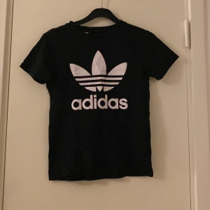 Svart Adidas t-shirt med logga - Svart t-shirt från Adidas med stor vit trefoil-logga och text på bröstet. Klassisk rund hals och korta ärmar. Tillverkad i mjuk bomull som är skön mot huden. Perfekt för dig som gillar sportig och enkel stil.