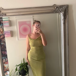 Ljusgrön långklänning med snörning - Säljer en ljusgrön långklänning med smala axelband och snygg draperad halsringning. Klänningen har snörning i ryggen och är figurnära med ett glansigt material som ger en lyxig känsla. Perfekt för dig som vill sticka ut med en cool och trendig look. Aldrig använd 