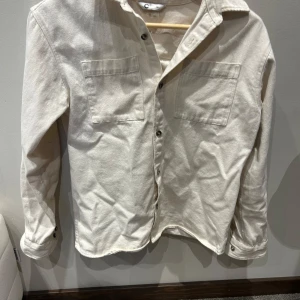 Beige overshirt i bomull från Cubus  - Stilren beige overshirt i bomull med två stora bröstfickor och knäppning framtill. Skjortan har klassisk krage och långa ärmar med knappar vid manschetterna. Perfekt att slänga över en t-shirt för en avslappnad look.