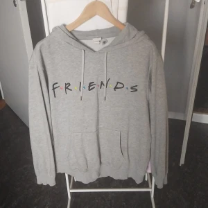 Grå Friends hoodie från H&M - Grå hoodie från H&M med Friends-tryck framtill och klassisk känguruficka. Tröjan har huva med snörning och långa ärmar. Perfekt för dig som älskar tv-serien Friends och vill ha en chill och snygg look.