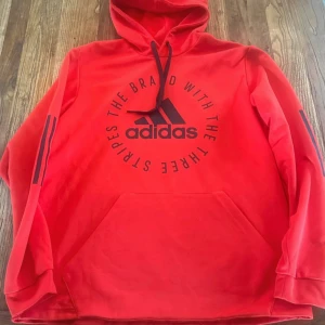 Röd Adidas hoodie med tryck - Säljer en röd hoodie från Adidas med svart logga och text på bröstet. Tröjan har huva med snörning, känguruficka och svarta ränder på ärmarna. Materialet är mjuk bomull/polyester och modellen är normal passform med långa ärmar. Perfekt för chill eller träning. Har tyvärr några små fläckar men inget man tänker på. 