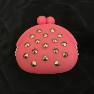 Rosa plånbok med nitar - Söt liten rosa plånbok i rund form med silvriga nitar på framsidan. Gjord i mjukt syntetmaterial och har en kulformad knäppning upptill. Perfekt för mynt eller småsaker och sticker verkligen ut i väskan.