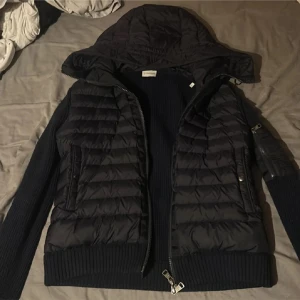 Svart Moncler pufferjacka med huva - Svart pufferjacka från Moncler med stickade ärmar och sidor, quiltade partier framtill och baktill samt stor huva. Jackan har dragkedja, ribbade muddar och en klassisk Moncler-logga på ärmen. Perfekt för dig som vill ha en snygg och sportig look.