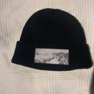 Svart ribbstickad mössa med patch - Limited edition Svart ribbstickad mössa från revenite med uppvikt kant och en stor rektangulär patch framtill. Patchen har ett svartvitt motiv av en person i vinterlandskap med berg i bakgrunden. Perfekt för dig som gillar stilrena accessoarer med unik detalj.