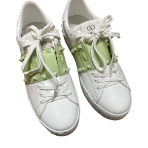 Valentino Garavani Rockstud sneakers - Snygga vita Valentino Garavani Rockstud sneakers med ljusgrönt band och ikoniska nitar. Skorna har vit snörning, chunky sula med svarta stänk och coolt rutmönster undertill. Tillverkade i skinn och perfekta för dig som vill sticka ut med stil.